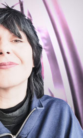 Petras Gesicht ist zur Hälfte zu sehen. Sie lächelt vor einem verschwommenen pinken Hintergrund. Sie hat einen schwarzen Mullet und trägt eine dunkelblaue Jacke.