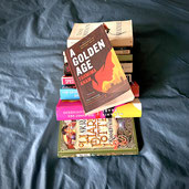 pile of books on grey material with a golden age by tahmima anam on top