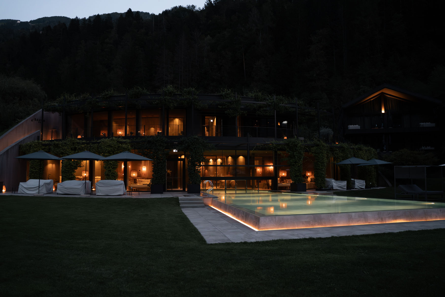 Nächtliche Außenansicht des luxuriösen Spa-Resorts mit beleuchtetem Pool, warmem Innenlicht und moderner Architektur vor bewaldeter Bergkulisse.