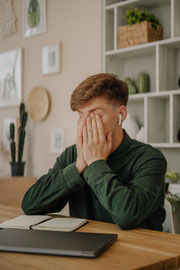 Ein rotblonder, weisser, junger Mann hält sich mit den Handinnenflächen die Augen zu und sitz an einem Schreibtisch mit einem Laptop. Der Laptop ist geschlossen. Im Hintergrund sieht man ein Bücherregal und Bilder an der Wand.