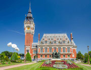 Façade en briques rouges de l'Hôtel de Ville de Calais et son beffroi classé à l'UNESCO, entourés d'un jardin fleuri sous un ciel bleu. Une idée de visite incontournable lors de votre séjour dans notre gîte sur la Côte d'Opale.