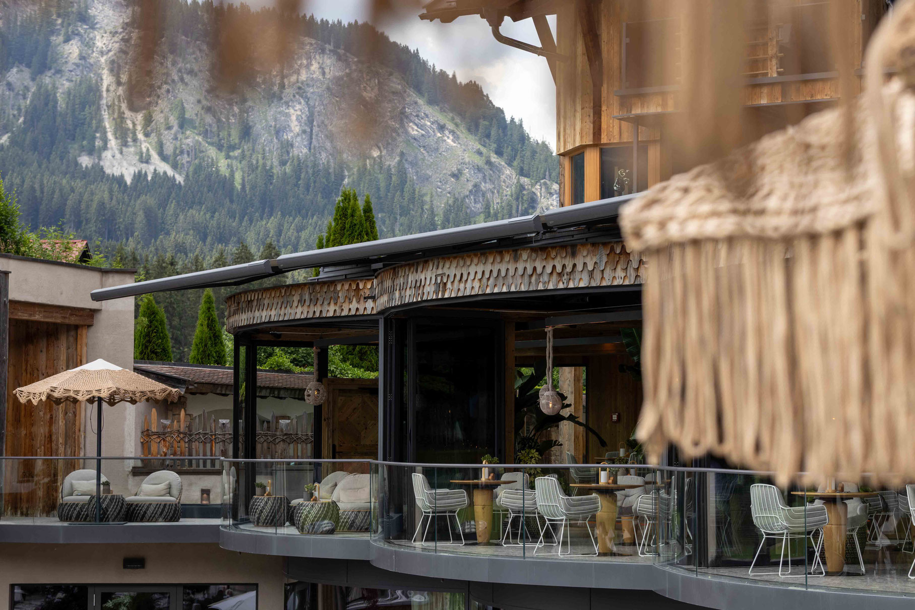Außenansicht eines modernen Hotels mit großzügiger Terrasse und raumhohen Glasfronten. Helle Sitzgruppen, natürliche Holzdetails und dekorative Sonnenschirme fügen sich harmonisch in die alpine Landschaft mit bewaldeten Berghängen im Hintergrund ein.
