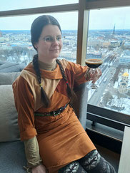 Girl with dark brown plaits and brownn dress holding a cocktail with city view in background in Riga's Skyline Bar
