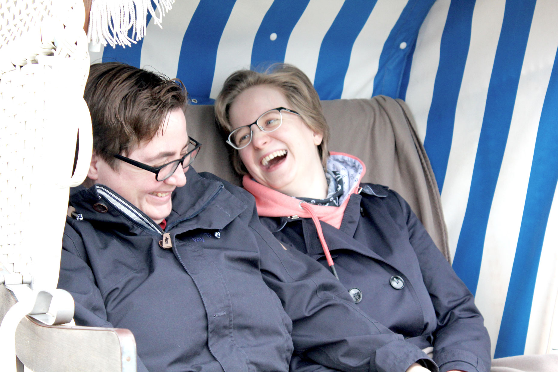 Beide Bräute, in Allwetterjacken im Strandkorb lachen aus vollem Herzen. Überraschungstrauung am Strand im November mit Constanze Marienfeld.