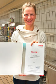 Teilnehmerin hält ihr Lehrabschlusszeugnis als Köchin in der Hand, im Küchenumfeld