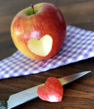 Roter Apfel, es ist ein Stück in Form eines Herzens herausgeschnitten. Das Stück liegt davor