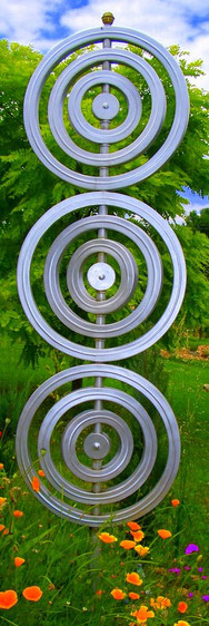 Vertikale Metallskulptur aus drei übereinander angeordneten Kreisformen, die das Logo der Psychotherapeutin darstellen, im Garten vor grünem Laub und Blumen.
