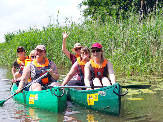 Foto von zwei Kanus mit jeweils drei Frauen mit Paddeln auf der Aller vor einem schilfbestandenen Ufer.