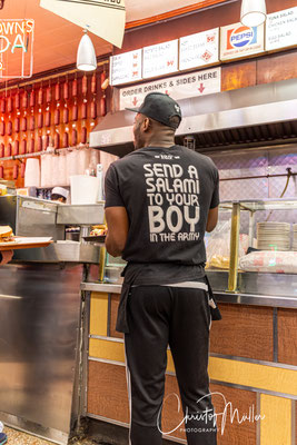 A bustling New York deli where speed, slogans, and nostalgia blend into a culture of performance and fast consumption—worlds apart from the quiet rhythms of rural life.