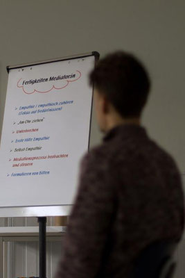Eine Person steht mit dem Rücken zum Betrachtenden vor einem Flipchart, auf dem erklärt wird, welche Fähigkeiten Mediatior:innen haben sollten.