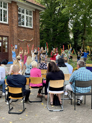 Kinder der Schule Strom führen mit bunten Boomwhackers eine Tanzeinlage auf. Alle Kinder halten die Boomwhackers nach oben. Eltern sitzen davor und schauen zu.