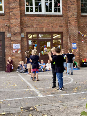 Kinder der zweiten Klasse führen einen Tanz vor, stehen zu zweit zusammen und klatschen sich mit beiden Händen ab. Im Hintergrund sitzen Schüler:innen und die Schulleiterin der Schule.