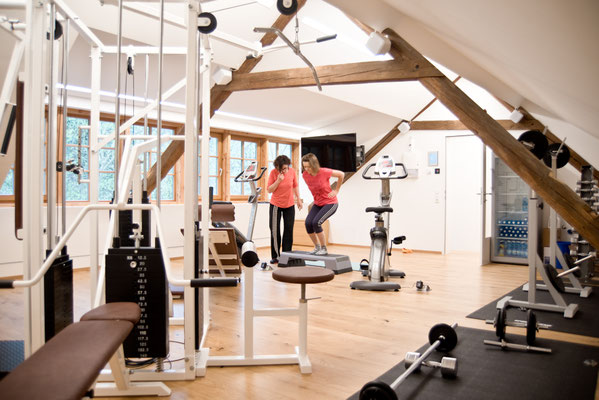 verschiedene Fitnessgeräte und im Hintergrund eine Physiotherapeutin die eine Patientin instruiert