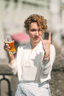 Braut in heller Kleidung hält ein Bierglas und zeigt eine selbstbewusste Geste, urbane Hochzeitsfotografie in Villach.