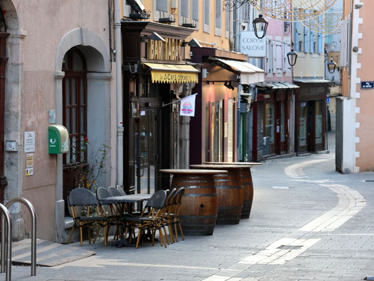 Sehenswürdigkeiten Gap: Blick in eine der Gassen in der Altstadt von Gap. Weinfässer sind vor einer Bar aufgestellt und werden als Tische genutzt. Eisenlampen hängen über der Gasse.