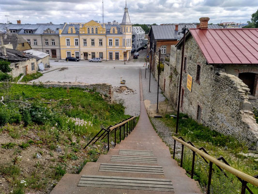 vorn ein schön restauriertes  Haus, links und rechts verfallende Gebäude, dazwischen eine moderne Treppe hinauf zur Burg 