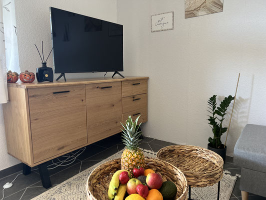 Fernsehecke der Ferienwohnung „Auszeit Bergliebe“ mit modernem Flachbild-TV auf einem stilvollen Holzsideboard. Im Vordergrund ein geflochtener Tisch mit frischem Obst – perfekt für entspannte Urlaubsabende in gemütlicher Atmosphäre.