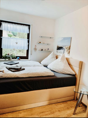 Helles Schlafzimmer der Ferienwohnung „Auszeit am See“ mit komfortablem Doppelbett, großen Kissen und liebevoll gefalteten Handtüchern. Ein Fenster mit Blick ins Grüne und dekorative Elemente schaffen eine freundliche und erholsame Atmosphäre.