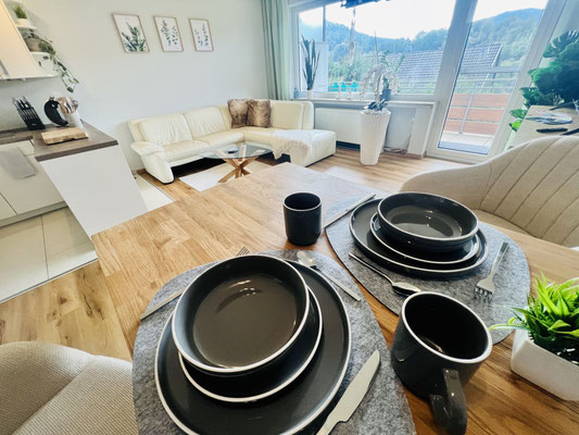 Einladend gedeckter Esstisch in der Ferienwohnung „Auszeit Niedersfeld“ mit stilvollem, schwarzem Geschirr und Ausblick ins gemütliche Wohnzimmer mit heller Eckcouch. Große Fenster sorgen für viel Licht und eine freundliche Atmosphäre.