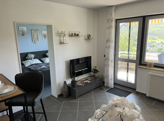 Heller Wohnbereich der Ferienwohnung „Auszeit Bergliebe“ mit Zugang zum Balkon, kleinem TV-Bereich und liebevoll dekorierten Wandregalen. Durch die offene Tür sichtbar: das gemütliche Schlafzimmer – alles harmonisch und einladend gestaltet.