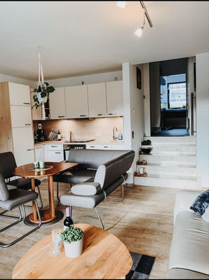 Offener Wohn- und Essbereich der Ferienwohnung „Auszeit am See“ mit moderner Küche, gemütlicher Sitzecke und Esstisch. Warme Holztöne, liebevolle Details und eine kleine Treppe zum oberen Bereich verleihen der Wohnung Charme und Wohnlichkeit.