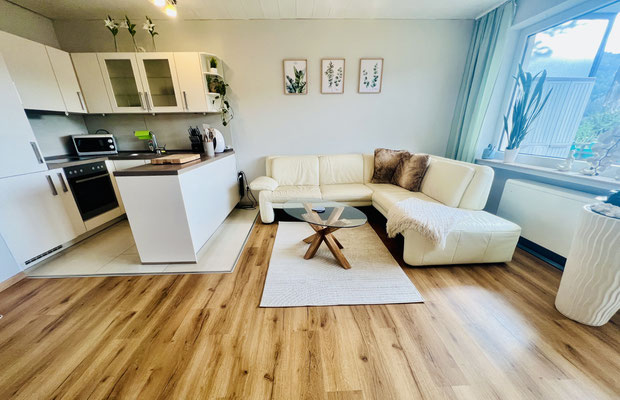 Heller Wohnbereich der Ferienwohnung „Auszeit Niedersfeld“ mit gemütlicher Eckcouch, modernem Couchtisch und stilvoller Dekoration. Die offene Küche und der Holzfußboden schaffen eine freundliche, einladende Atmosphäre zum Wohlfühlen.