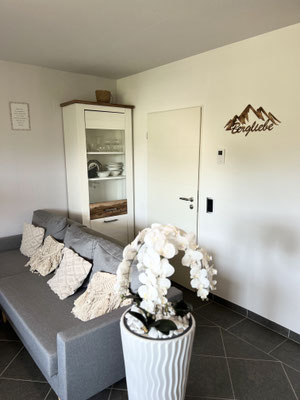 Gemütliche Sitzecke in der Ferienwohnung „Auszeit Bergliebe“ mit grauem Sofa, dekorativen Kissen und stilvoller Orchidee. An der Wand das passende Holzschild „Bergliebe“ – liebevoll gestaltet für entspannte Auszeiten mit alpinem Flair.