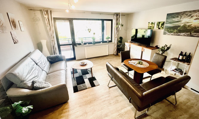 Wohnzimmer der Ferienwohnung „Auszeit am See“ mit zwei gemütlichen Sofas, Essbereich und Zugang zum Balkon. Große Fenster lassen viel Licht herein, stilvolle Deko, Pflanzen und ein Flachbild-TV schaffen eine wohnliche und entspannte Atmosphäre.