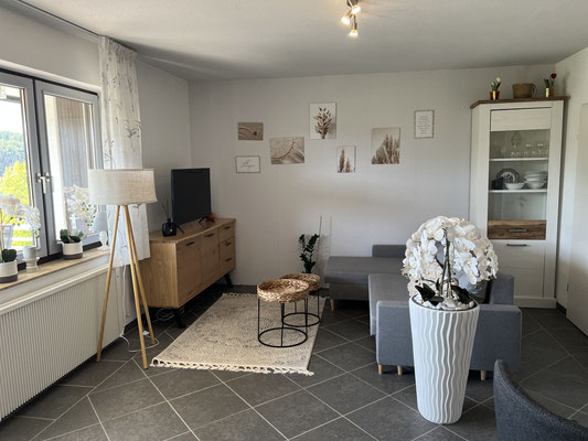 Wohnzimmer der Ferienwohnung „Auszeit Bergliebe“ mit grauer Eckcouch, TV-Bereich und stilvoller Einrichtung. Eine große Orchidee, Naturmotive an der Wand und gemütliche Beleuchtung schaffen eine harmonische und einladende Wohlfühlatmosphäre.
