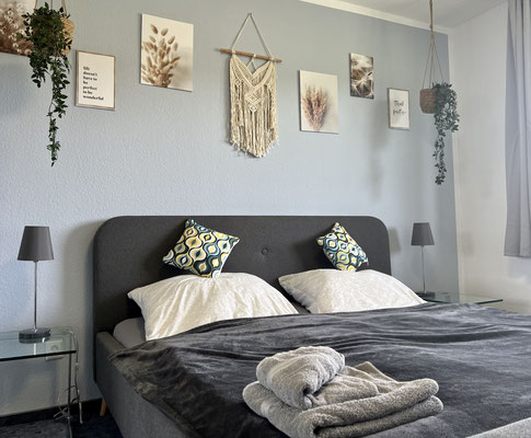 Schlafzimmer der Ferienwohnung „Auszeit Bergliebe“ mit gemütlichem Doppelbett, geschmackvoller Wandgestaltung mit Naturmotiven und Makramee sowie liebevoll gefalteten Handtüchern – ideal für erholsame Nächte in stilvollem Ambiente.