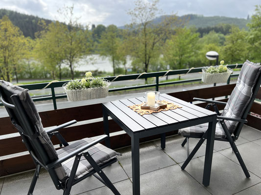 Balkon der Ferienwohnung „Auszeit am See“ mit Sitzgruppe und traumhaftem Blick auf den Hillebachsee und die umliegende Natur. Blumenkästen, gemütliche Hochlehner und ein dekorierter Tisch laden zum Verweilen und Genießen ein.