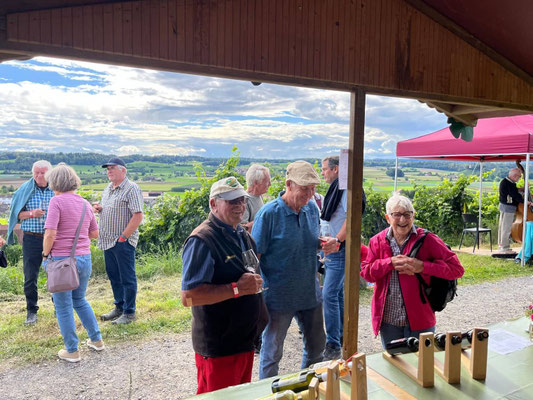 Aus einem Fenster heraus sieht man zwei lachende Männer und eine Frau, die mit einem Weinglas in der Hand auf einem Weg im Rebberg stehen
