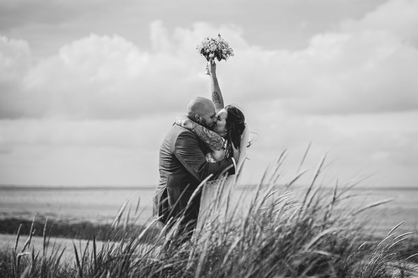 Brautpaar küsst sich in den Dünen in Hooksiel an der Nordsee und sie hält ihren Brautstrauß in die Luft.
