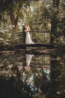 Hochzeitspaar steht auf einer Brücke und spiegelt sich im Bach.
