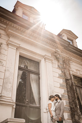 Brautpaar steht im Sonnenlicht an einem Gebäude.