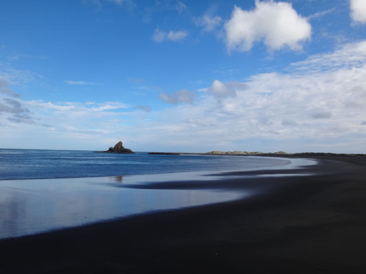 Eine spiegelglatte, feuchte Sandoberfläche und ein entfernter Leuchtturm am Whatipu Beach