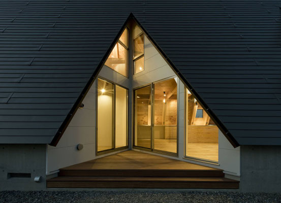 岐阜県にある大きな三角屋根が特徴的なモダンな注文住宅「合掌の家」の夜景。急勾配の屋根の開口部から室内の暖かな照明が漏れ、ウッドデッキとガラス張りのリビングが輝く美しい外観。