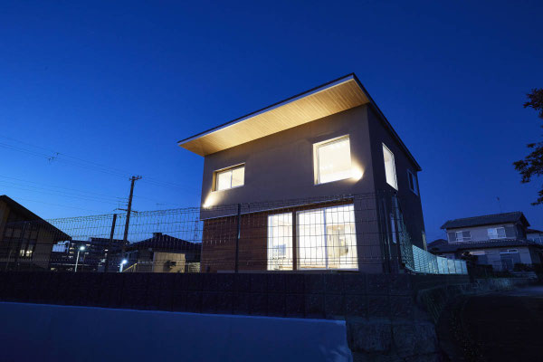 兵庫県の注文住宅の夜の外観。ブラウンの外壁と、明るく照らされた木目調の深い軒天。1階の大きな掃き出し窓と2階の窓から室内灯が漏れ、外壁のスポットライトが建物を立体的に浮かび上がらせている様子。