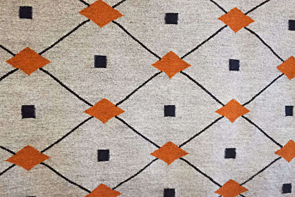 Fragment d'un grand tapis kilim tissé à la main en laine, au fond gris clair, les fines rayures noires en diagonal, les diamants oranges et petits carrés noirs. C'est un motif losange.