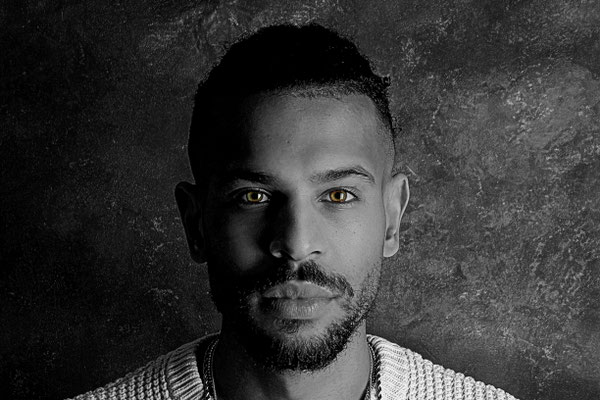 Schwarz-Weiß-Porträt eines Mannes mit markantem Blick, aufgenommen vor einem strukturierten, dunklen Hintergrund in einem Fotostudio.