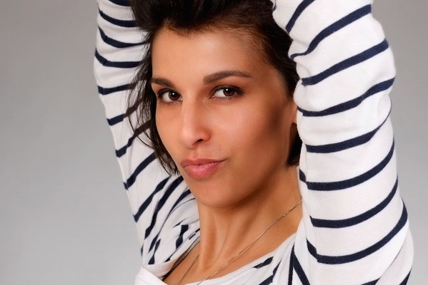 Professionelles Porträt einer Frau in gestreiftem Oberteil, die ihre Arme hinter dem Kopf verschränkt, aufgenommen vor einem neutralen Hintergrund in einem Fotostudio.