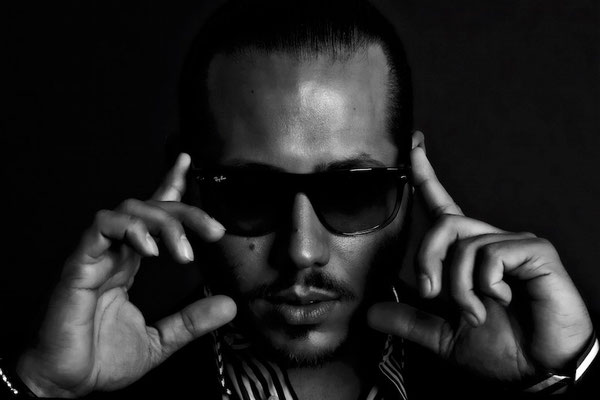 Schwarz-Weiß-Porträt eines Mannes mit Sonnenbrille, der seine Hände an die Brillengläser hält, aufgenommen vor einem dunklen Hintergrund in einem Fotostudio.