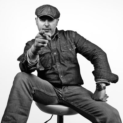 Schwarz-Weiß-Porträt eines Mannes in lässiger Pose, der auf einem Hocker sitzt und direkt in die Kamera zeigt, aufgenommen vor einem weißen Hintergrund in einem Fotostudio.