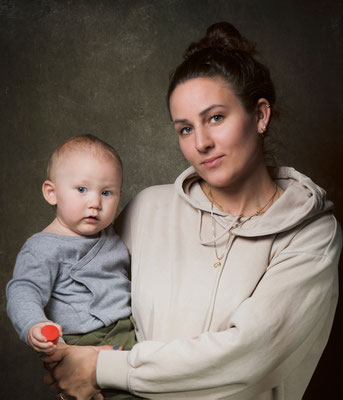Professionelles Porträt einer Frau, die ein Kleinkind hält, aufgenommen vor einem dunklen Hintergrund in einem Fotostudio.