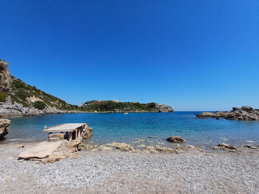 Auf der linken Seite des Fotos ist ein Steg zu sehen, der ins Meer führt. Im Vordergrund ist ein Kiesstrand zu sehen, dahinter das türkisblaue Meer mit Küste.