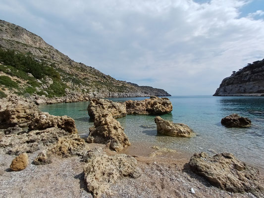 Im vorderen bereich des Fotos sieht man markante Felsformationen auf dem Stein und-KIesstrand. Dahinter ist das blaue Meer zu erkenne sowie der Eingang zur Bucht.