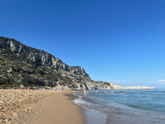Im Vordergrund ist der goldene Sandstrand zu sehen. Im rechten Teil ist das Meer und der blaue Himmel zu sehen. Auf der linke Seite ist der Beginn der Küste als größeren Ausschnitt zu sehen..