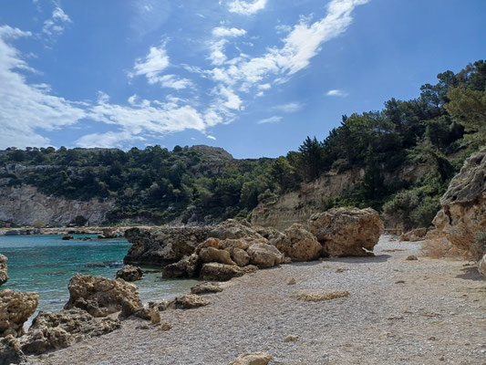 Im vorderen Bereich des Fotos ist der Kiesstrand zu sehen, dahinter ein größerer Felsen sowie das türkises Meerwasser und weitere Felsen im Hintergrund. Dahinter ist ein Pinienwald zu sehen.
