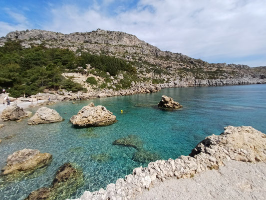 Auf dem Foto sieht man das türkisgesprenkelte Meer der Bucht sowie die markanten Felsformationen, die vom kristalklaren Wasser umgeben werden. Im Hintergrund ist die Küstenlandschaft zu sehen.