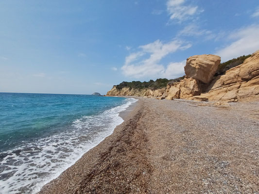 Auf der rechten Seite des Fotos ist ein Kiesstrand zu sehen mit großen Felsen. Auf der linken Seite ist das Meer zu sehen.
